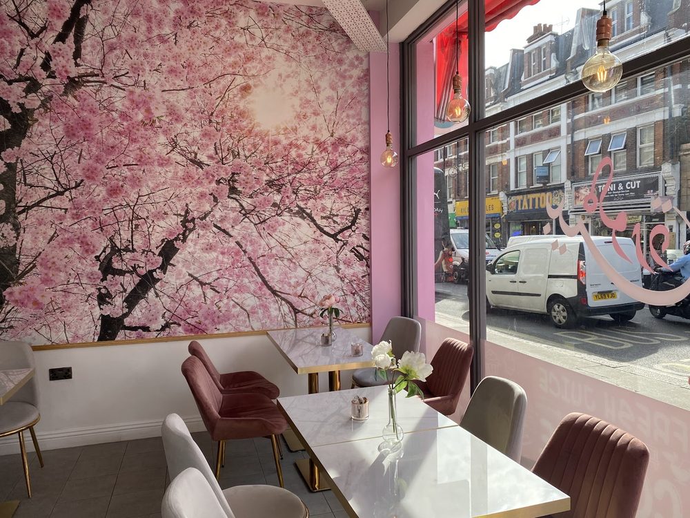 Window seating with marble tables and pink and grey chairs against a cherry-blossom wall mural, looking out onto Kilburn High Road.