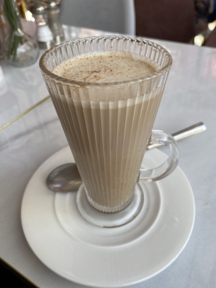 A tall fluted glass of dusted milky latte on a saucer with a long spoon.